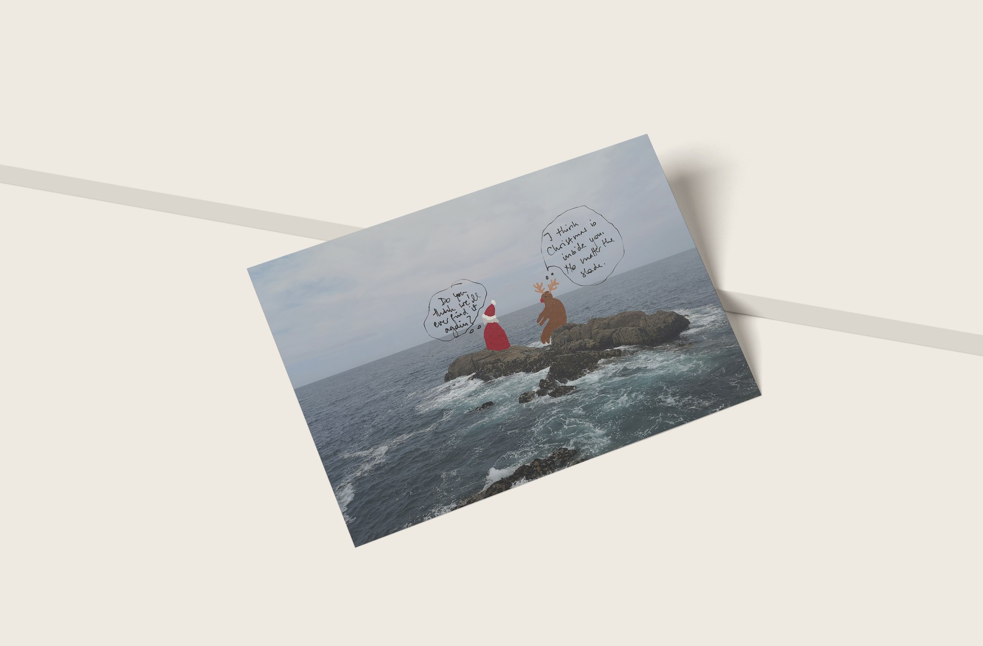 The postcard lying on a pale table across a white ribbon: a drawn Santa and reindeer on a rock in a grey sea