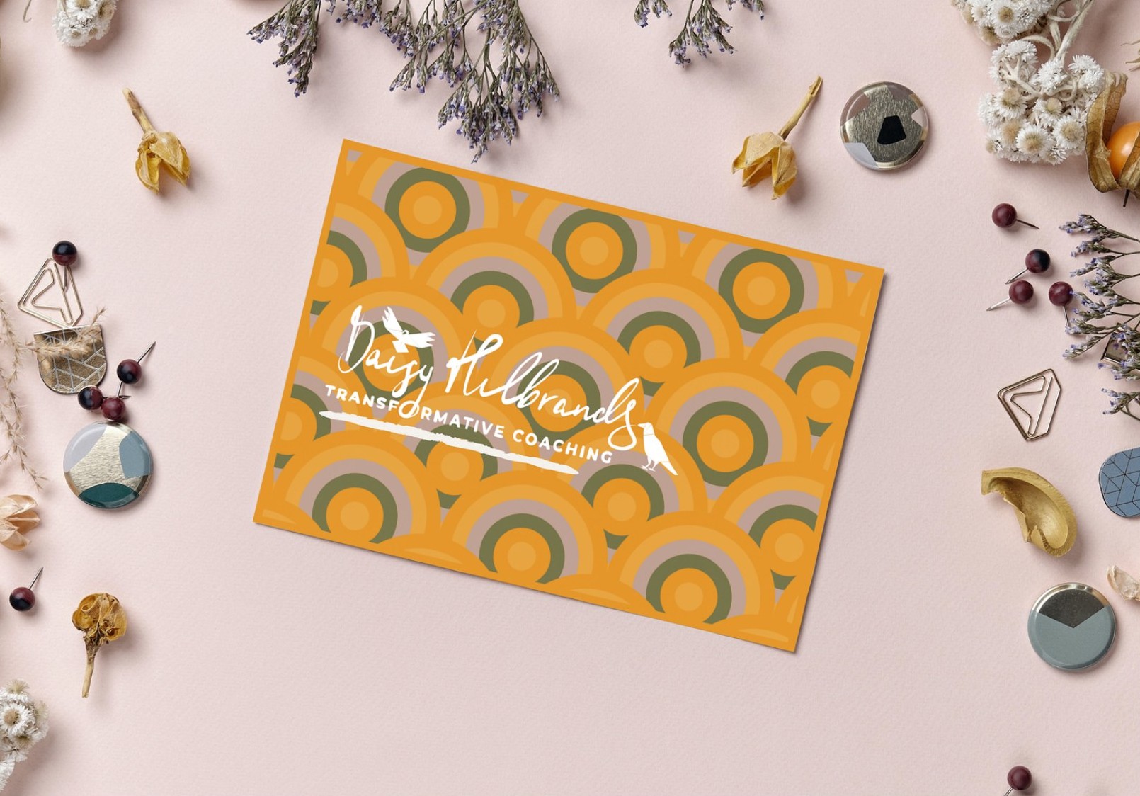 The orange postcard with her logo on the circle pattern, lying on a pink desk among dried flowers, pins and paper clips