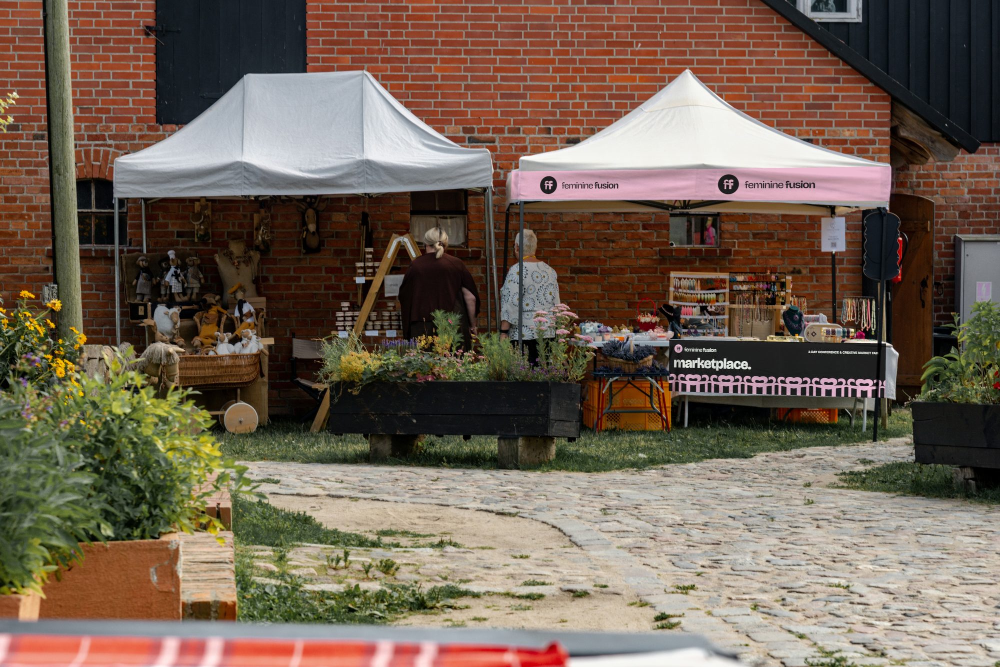 A market stall in front of a brick wall: the tent's valance printed pink with the logo twice, a black table banner marketplace. with the pink row along its bottom, jewellery on the table