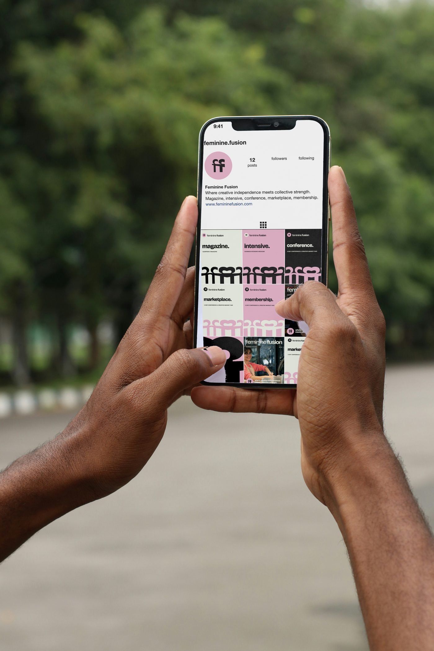 Two hands holding a phone with the feminine.fusion Instagram profile: the pink disc, the line, the grid of posts and covers