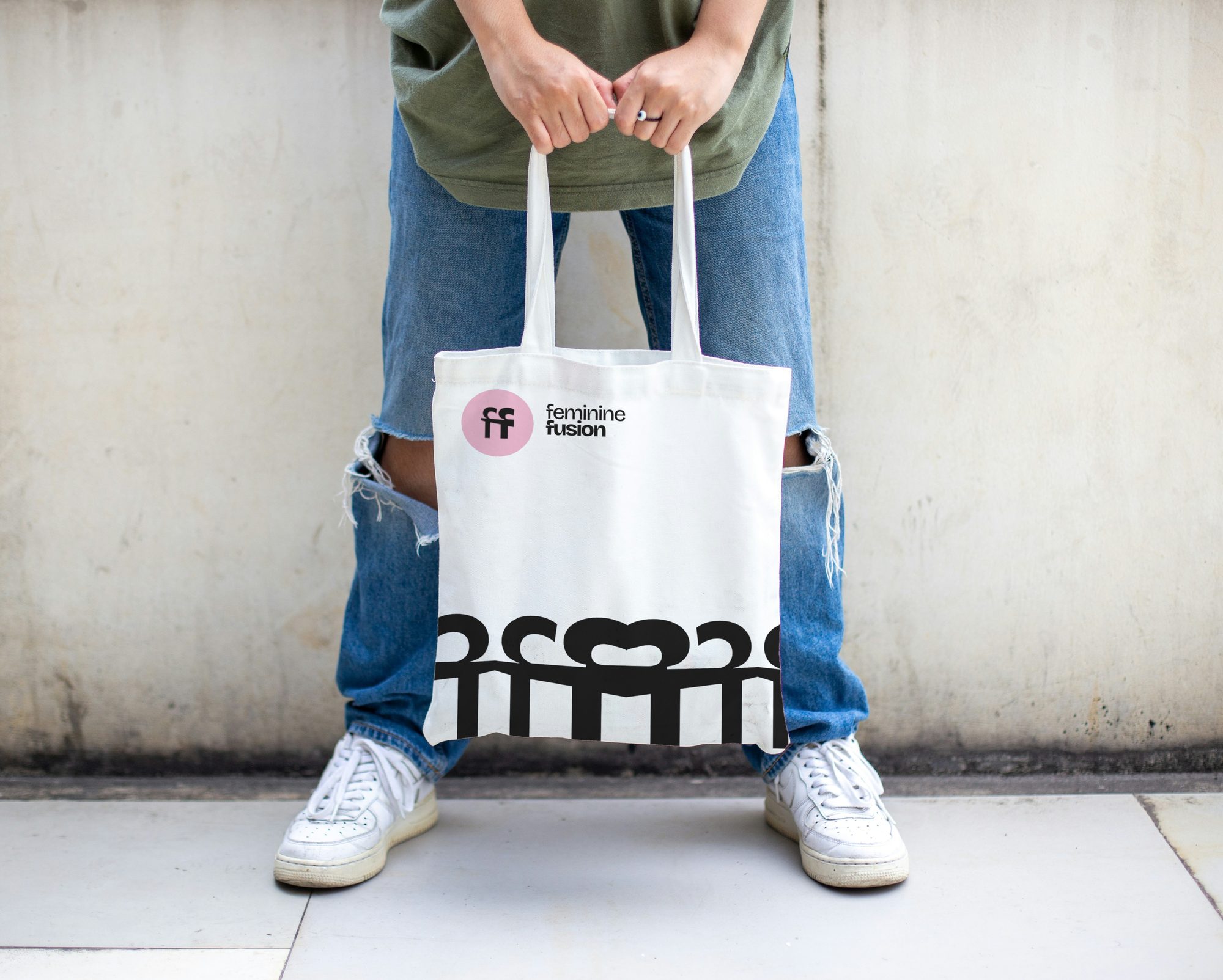 A white tote held in front of jeans: the pink disc and the stacked name top left, the black row of figures along its bottom