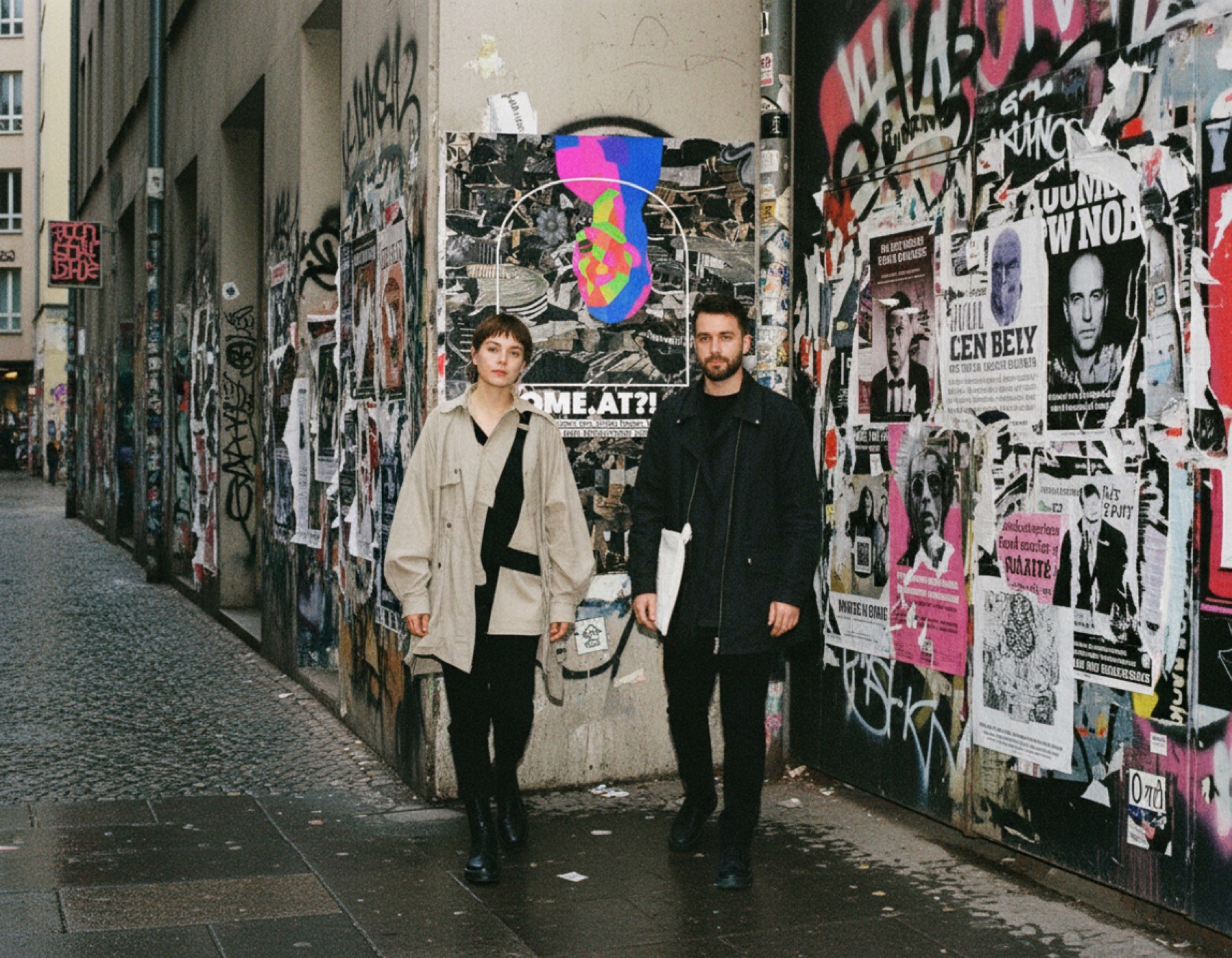 The poster on a street wall full of posters, two people walking past