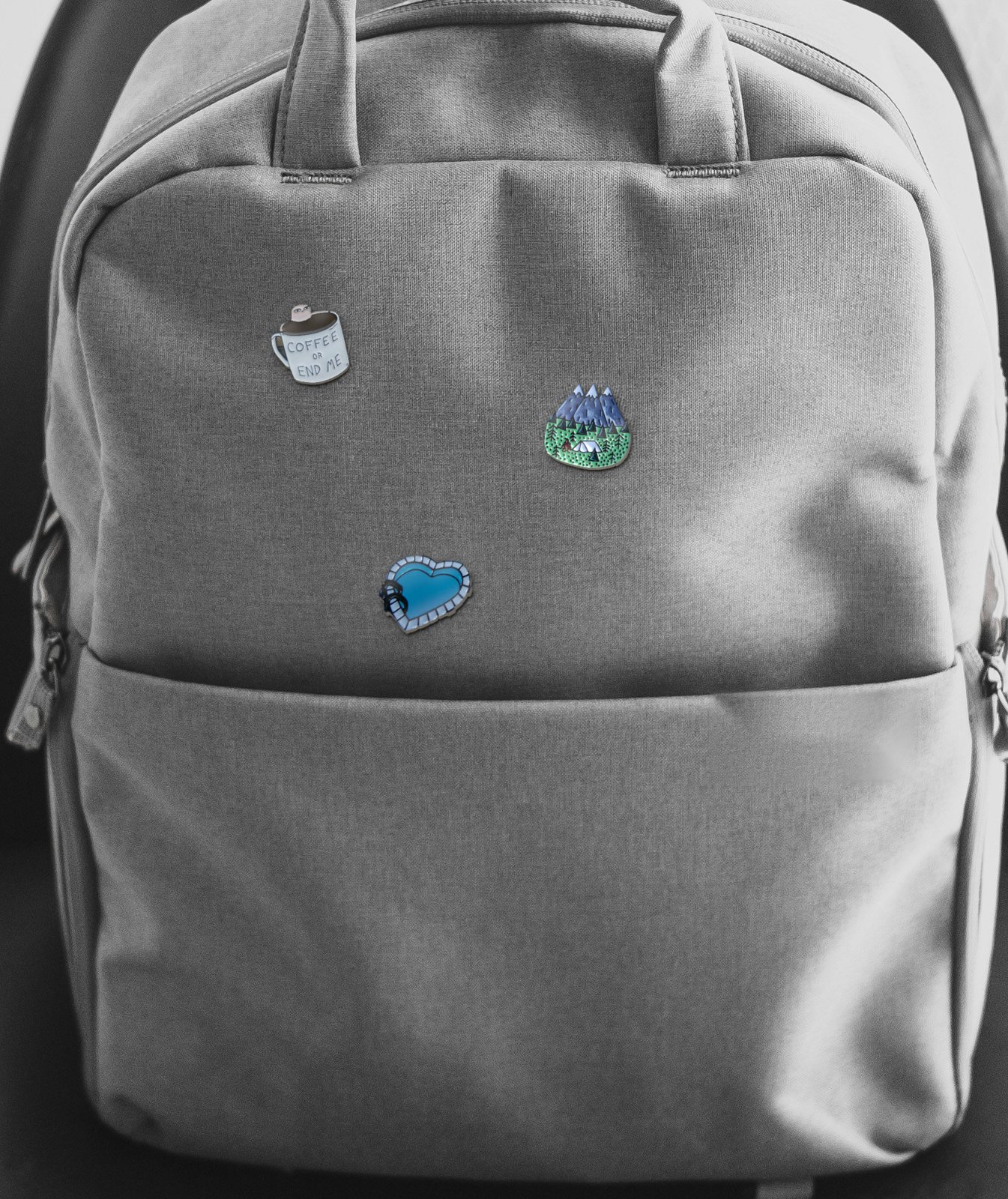 A plain grey backpack with three of the shop's pins on its front: a Coffee or End Me mug, a mountain tent and a blue heart