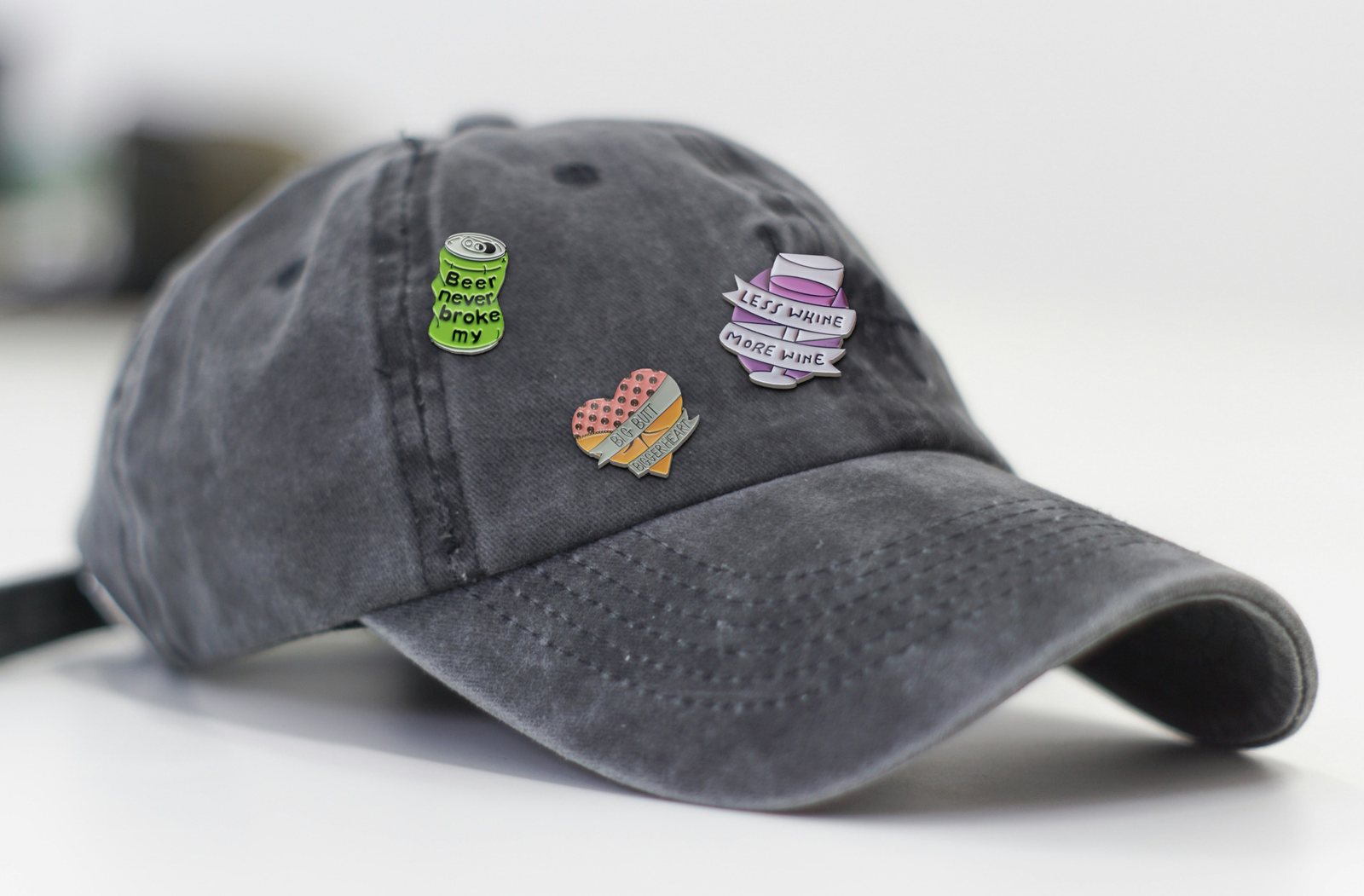 A washed grey cap with three pins on its front: a green can, a heart and a lilac banner