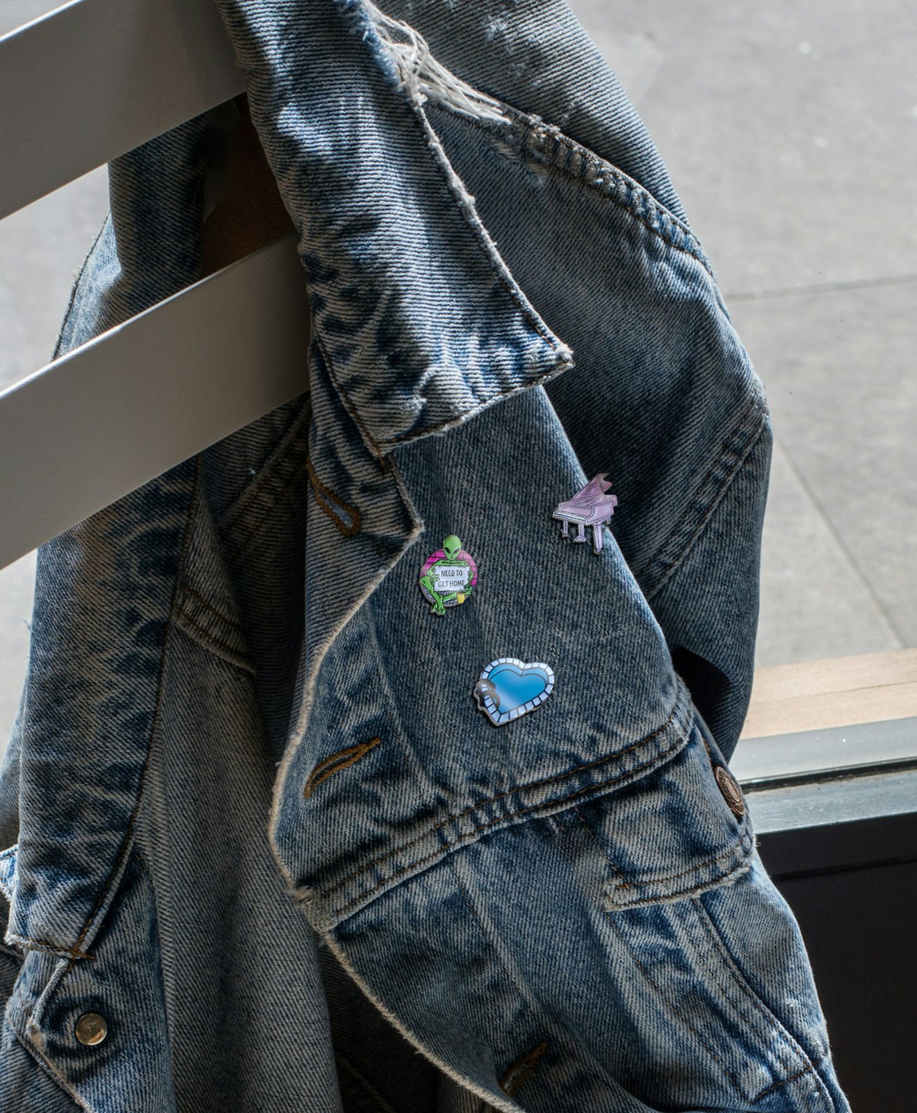 A denim jacket over a railing, three pins on its collar and chest: the green alien, the pink piano and the blue heart