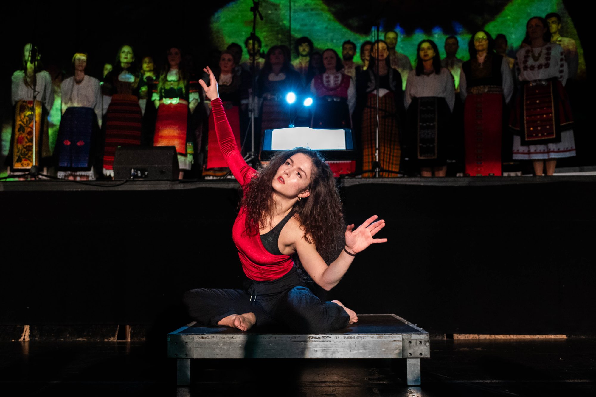 A dancer in red on a low platform, the choir behind her under a green projection