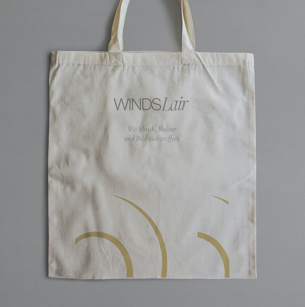 A natural cotton tote lying flat on grey: the grey Winds Lair wordmark, the line Wo Musik, Kultur und Bild sich treffen, and large gold arcs running off the bottom seam